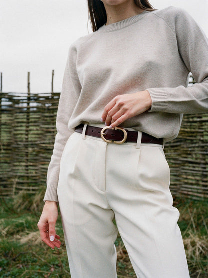 Womens leather belt in mahogany worn on the waist of cream trousers, brushed gold oval buckle