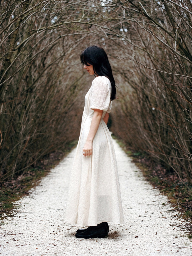 The same long dress in Candlelight, a warm milky off-white, worn full length under a bare hornbeam walk