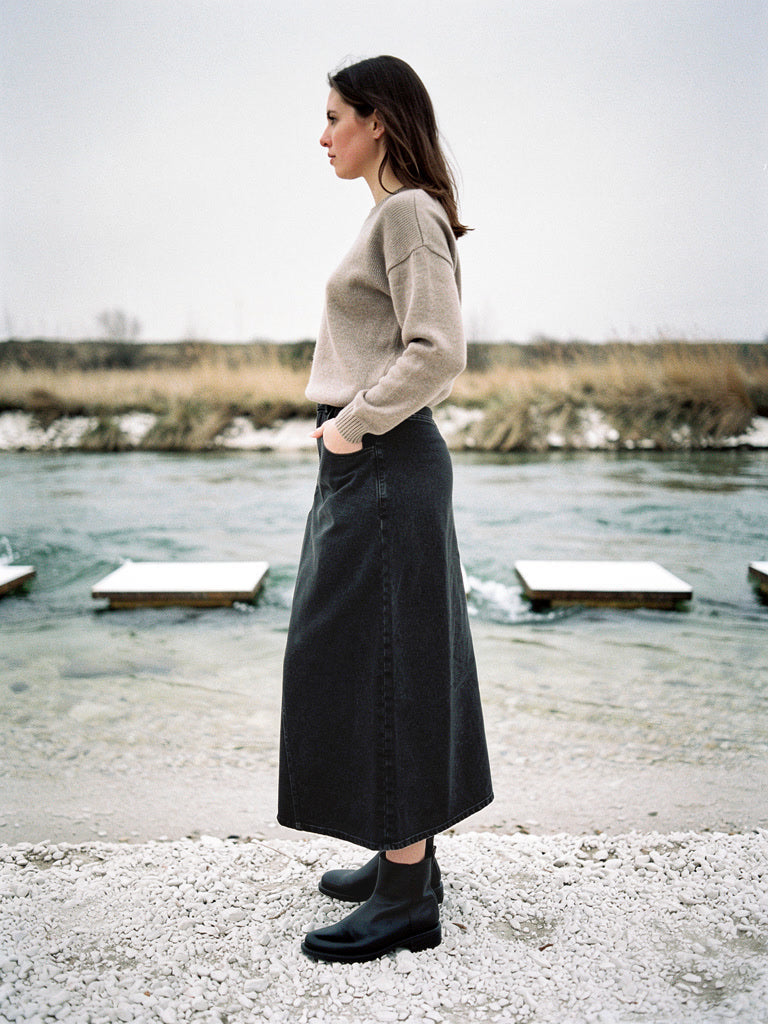 Long jean skirt in gunmetal washed charcoal denim worn in profile beside a shallow chalk river with stepping stones, the hem grazing the ankle