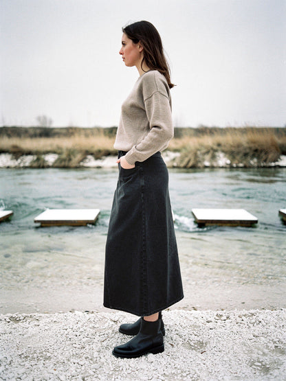 Long jean skirt in gunmetal washed charcoal denim worn in profile beside a shallow chalk river with stepping stones, the hem grazing the ankle