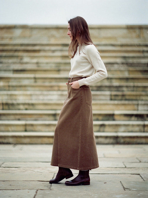 Long jean skirt in tanbark brown stretch denim worn with a cream jumper and flat boots, standing in an empty stone dry dock, the ankle length A-line hem falling unbroken to the boot