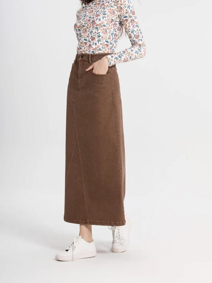 Long jean skirt in tanbark brown denim photographed three quarters on against a plain studio wall, one hand in the pocket, the hem level at the ankle