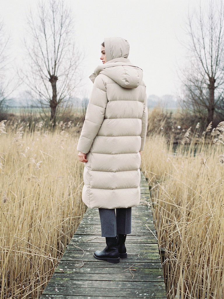 Woman wearing a long puffer coat in Parchment with the hood up on a fen boardwalk