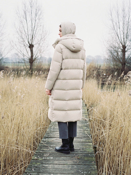 Woman wearing a long puffer coat in Parchment with the hood up on a fen boardwalk
