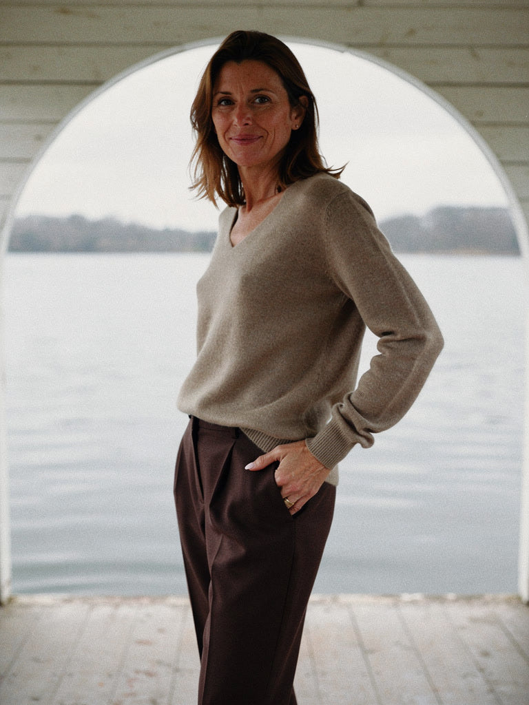 Woman wearing a Harlbury merino V-neck jumper in Driftwood inside a lakeside boathouse