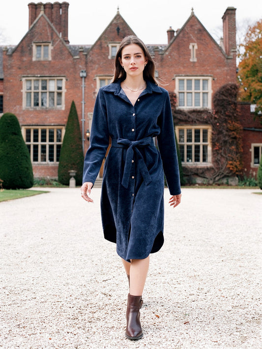 Womens midnight blue corduroy dress worn belted, walking on the gravel forecourt of a red brick Jacobean manor