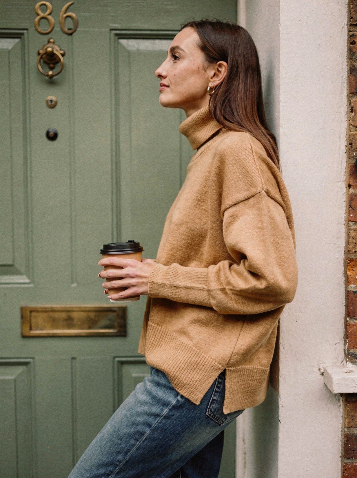 The camel oversized funnel neck jumper worn with jeans, side view showing the split hem