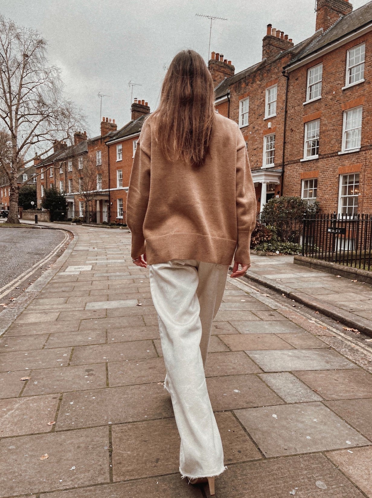 Back view of the camel oversized funnel neck jumper with split hem, worn with off-white wide-leg trousers on an English street