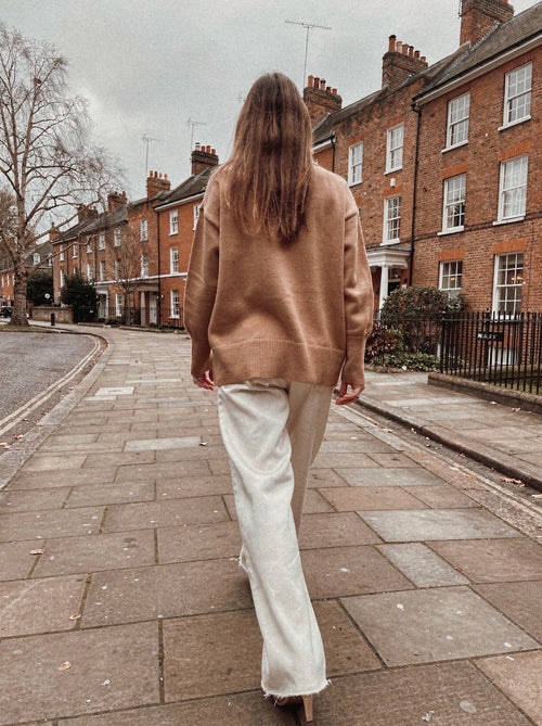 Back view of the camel oversized funnel neck jumper with split hem, worn with off-white wide-leg trousers on an English street