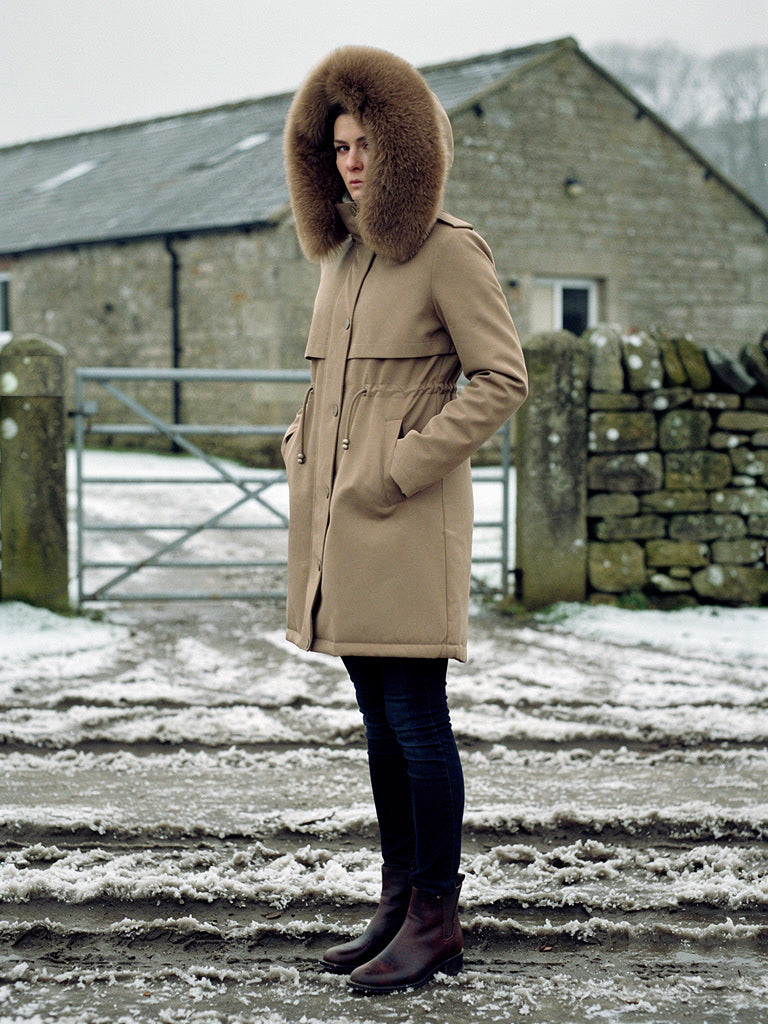 Womens parka coat in Sandstone with a deep faux fur trim hood, worn in a Yorkshire Dales farmyard