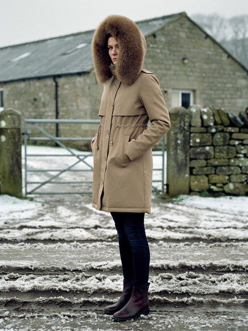 Womens parka coat in Sandstone with a deep faux fur trim hood, worn in a Yorkshire Dales farmyard