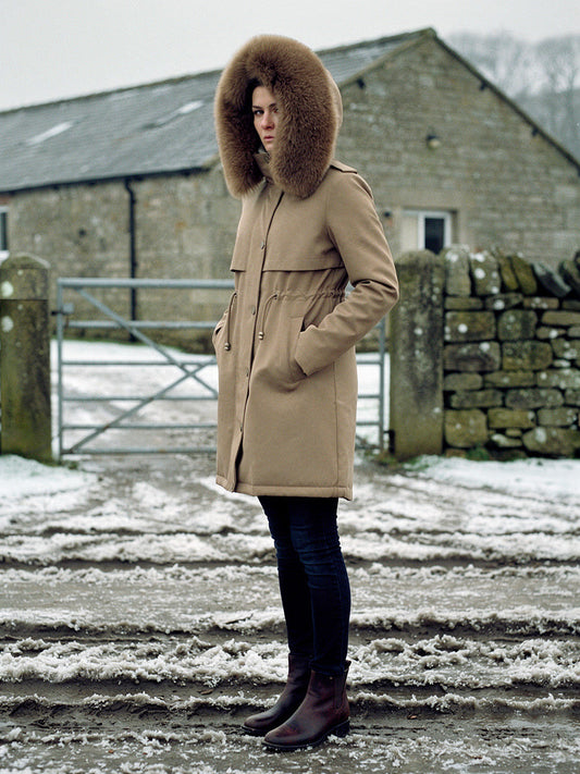Womens parka coat in Sandstone with a deep faux fur trim hood, worn in a Yorkshire Dales farmyard