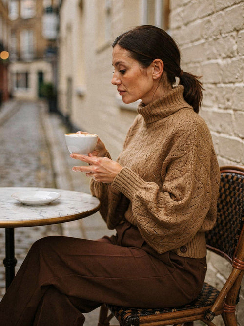 The camel pointelle roll neck jumper worn at a cafe table, three-quarter view