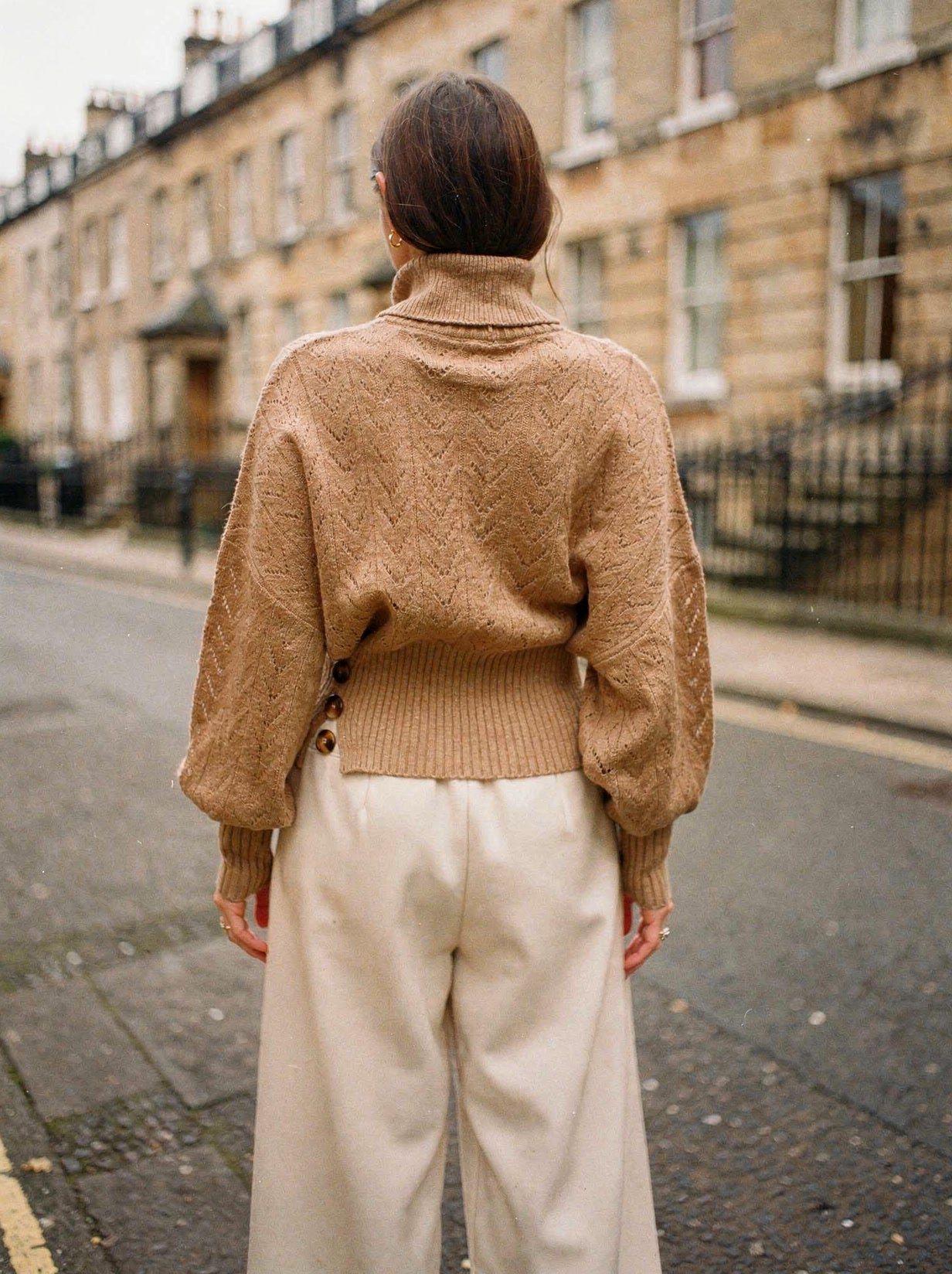Back view of the camel pointelle roll neck jumper with batwing sleeves, worn with cream wide-leg trousers