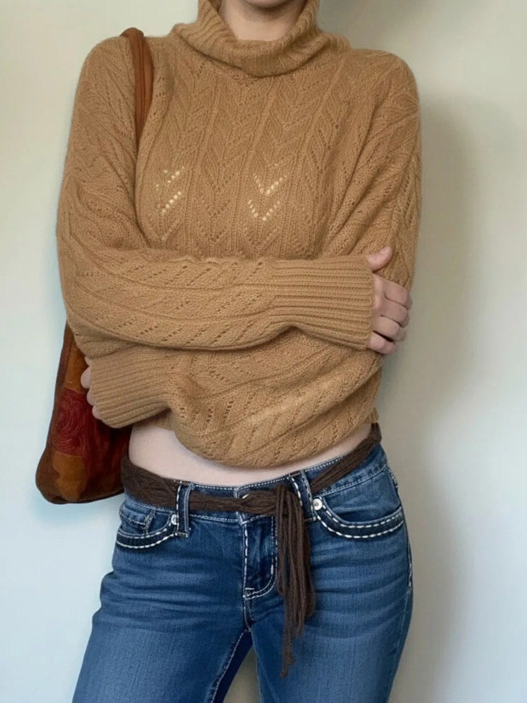 The camel pointelle roll neck jumper worn with arms crossed, showing the batwing sleeves