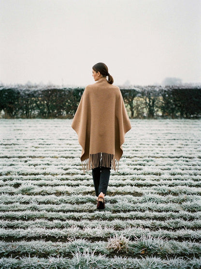 Woman walking away across a frosted ridge and furrow field, wearing the Hazel poncho cape open over her shoulders