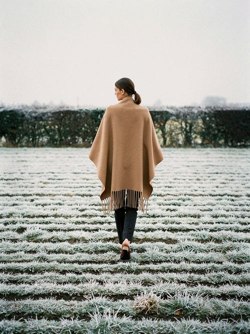 Woman walking away across a frosted ridge and furrow field, wearing the Hazel poncho cape open over her shoulders