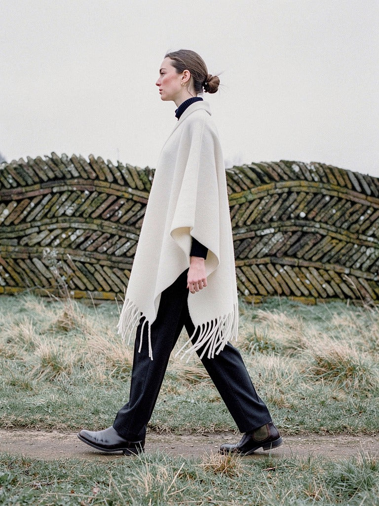 Woman walking in profile past a low serpentine brick wall, wearing the Muslin poncho cape