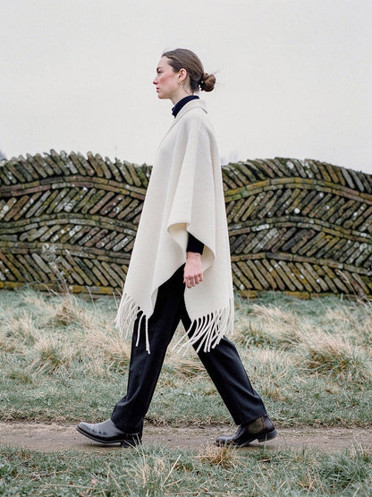 Woman walking in profile past a low serpentine brick wall, wearing the Muslin poncho cape