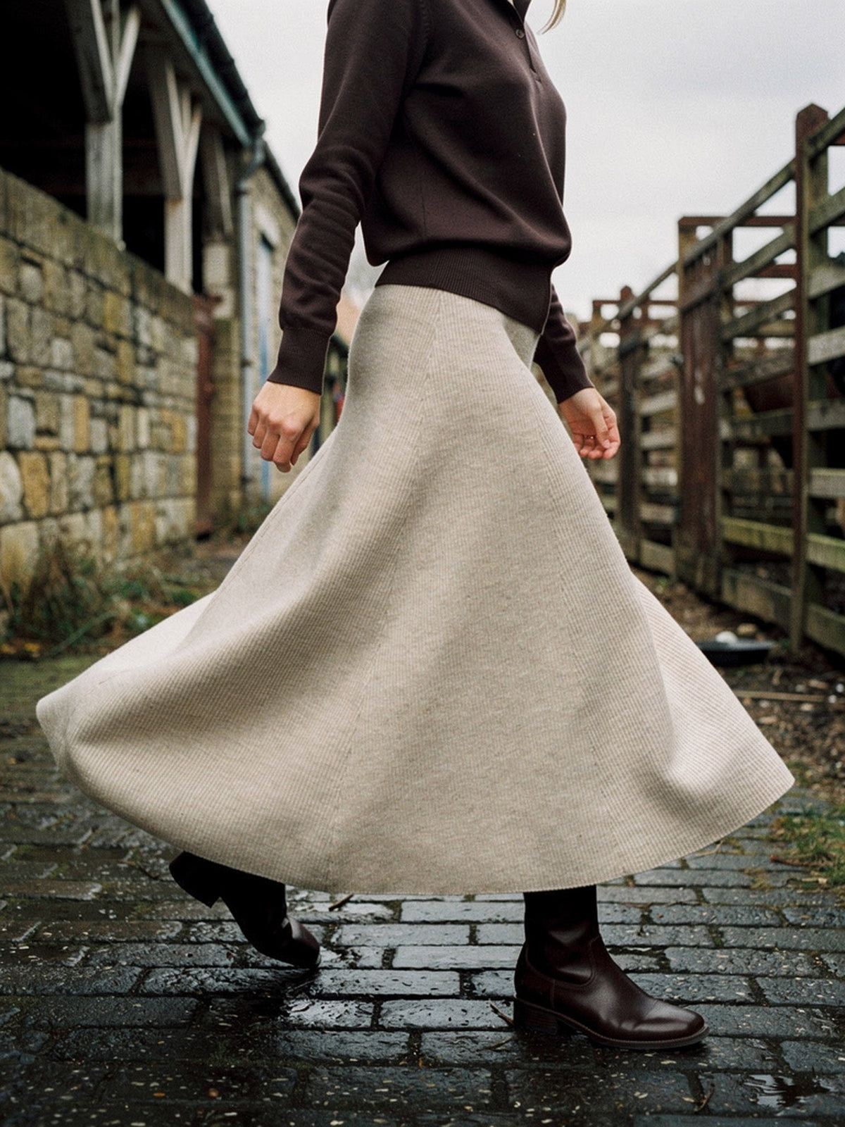 Womens ribbed midi skirt in porridge oatmeal knit, worn with a dark brown polo neck and flat boots, walking across the wet setts of an old cattle market so the full A-line sweep opens out