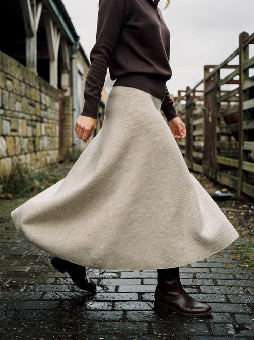 Womens ribbed midi skirt in porridge oatmeal knit, worn with a dark brown polo neck and flat boots, walking across the wet setts of an old cattle market so the full A-line sweep opens out