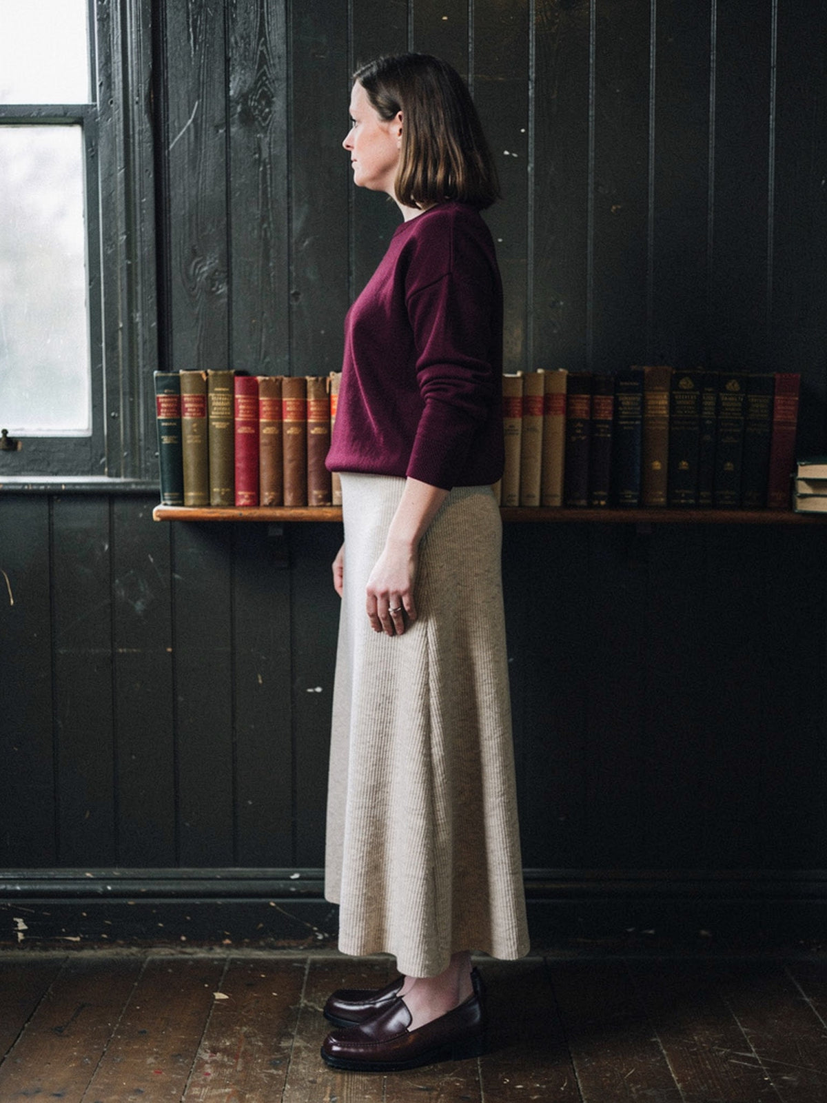 Side view of the porridge ribbed midi skirt in wear, the hem falling low on the calf, photographed in a panelled village reading room