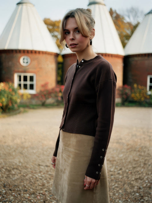 Womens cropped round-neck cardigan in Bitter Chocolate with silver press studs, worn in an oast house courtyard