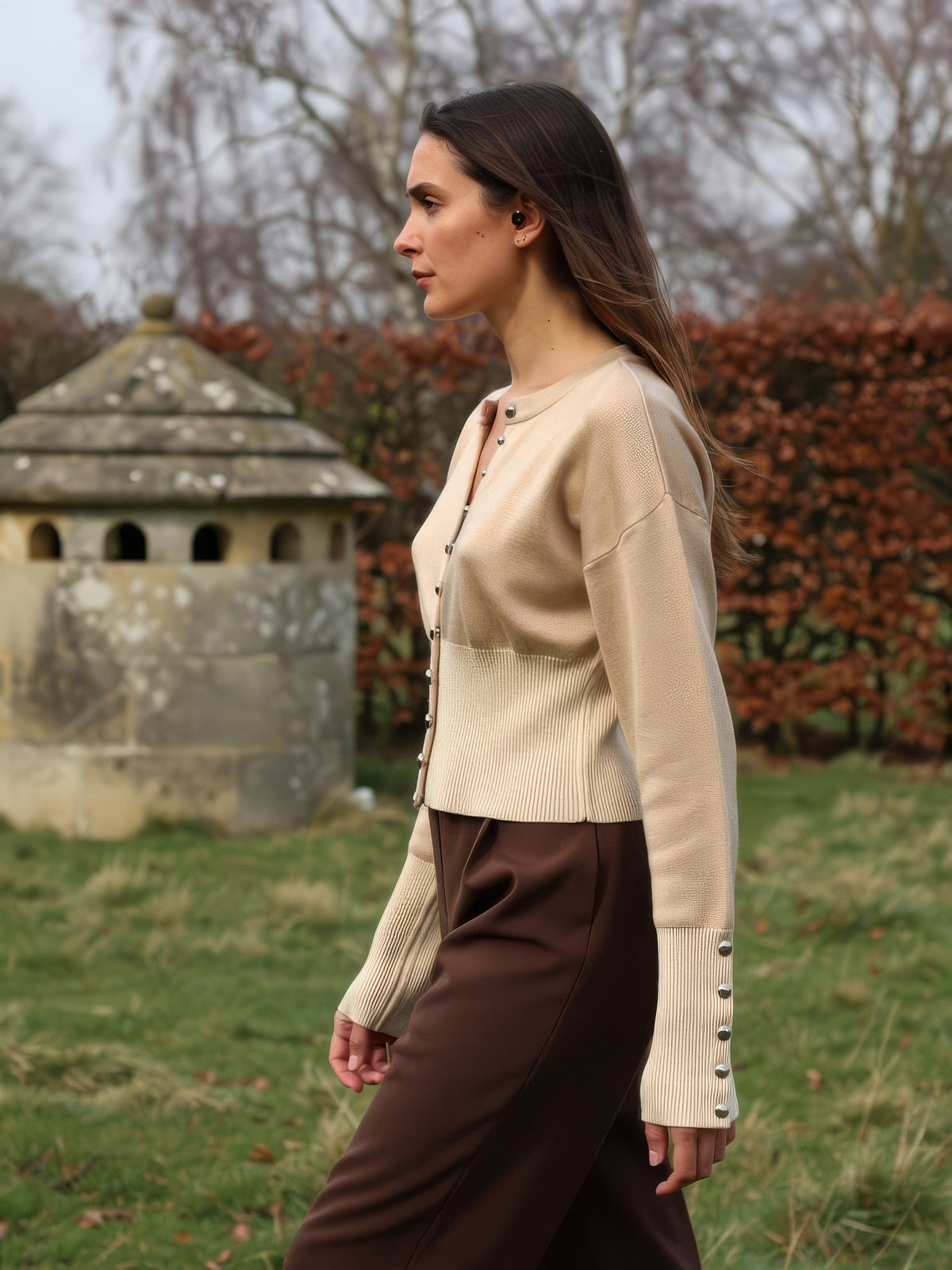 Womens cropped round-neck cardigan in Wheat worn in profile beside a stone dovecote