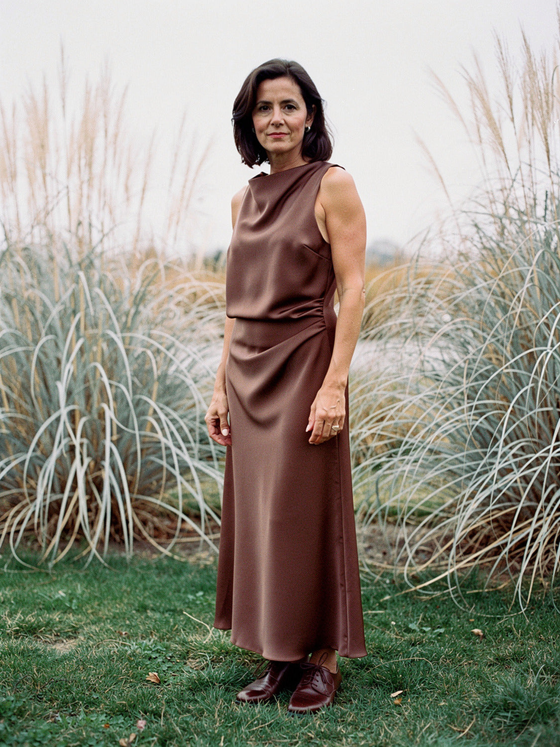 Woman wearing a Pecan brown satin maxi dress with a draped cowl neck in an autumn grass garden