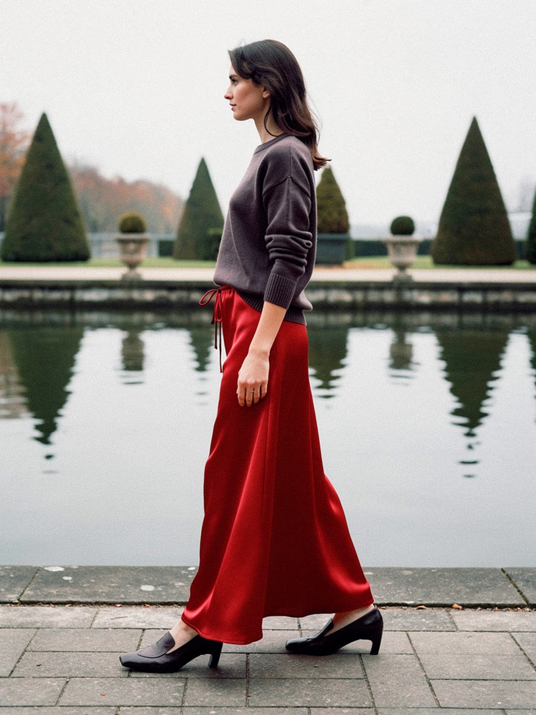 Woman walking in a poppy red satin maxi skirt beside a garden reflecting pool