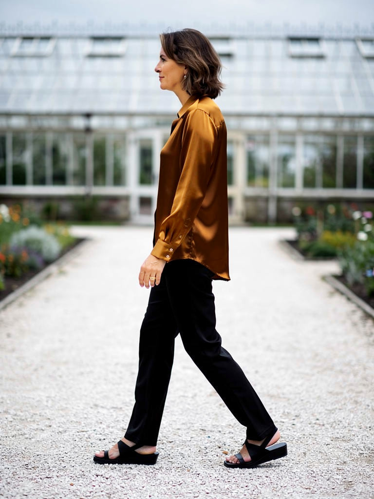 Woman walking in an amber satin shirt worn loose beside Victorian glasshouses