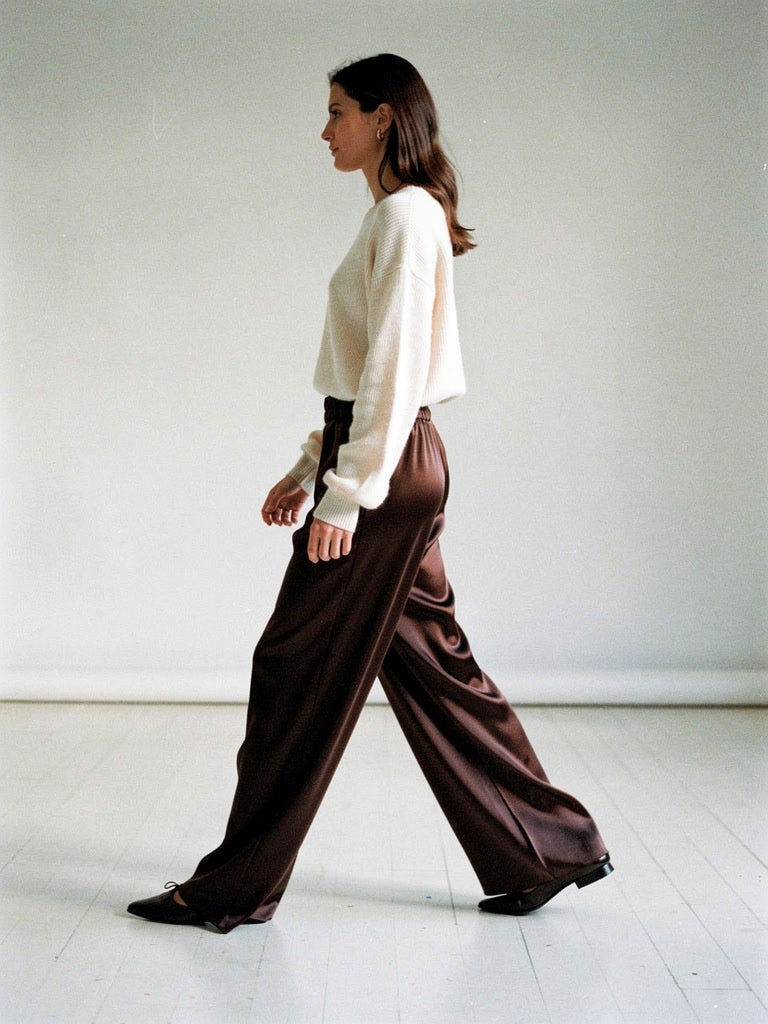 Side view of the chocolate satin trousers mid stride, the wide column leg falling to the floor