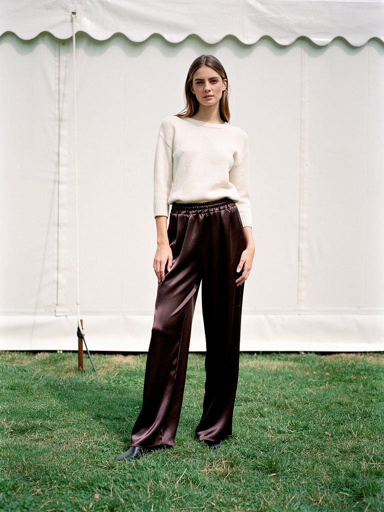 Woman in chocolate satin trousers with a wide column leg, standing on a lawn beside a marquee