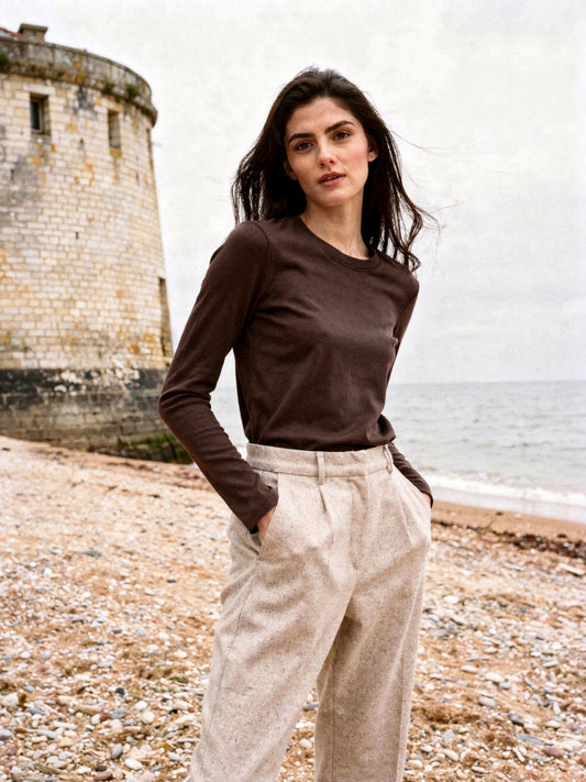 Woman on an English shingle beach wearing the chocolate silk blend top with a ribbed crew neck and oatmeal wool trousers