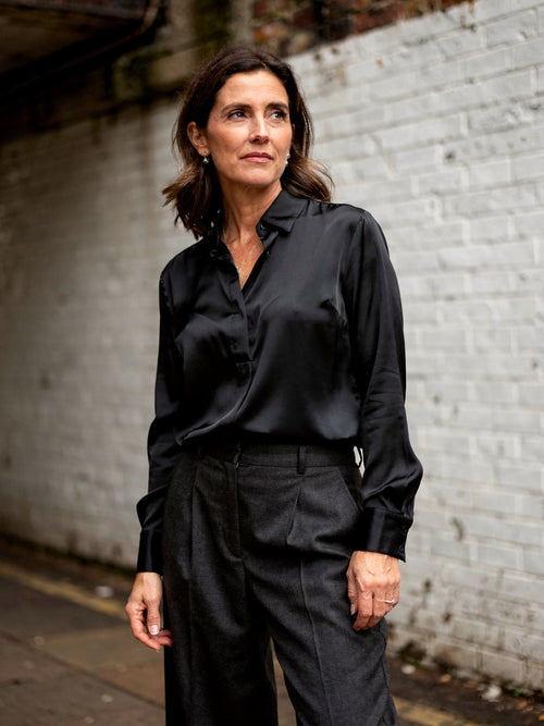 Woman wearing a black silk shirt tucked into charcoal wide leg trousers in front of a white glazed brick wall