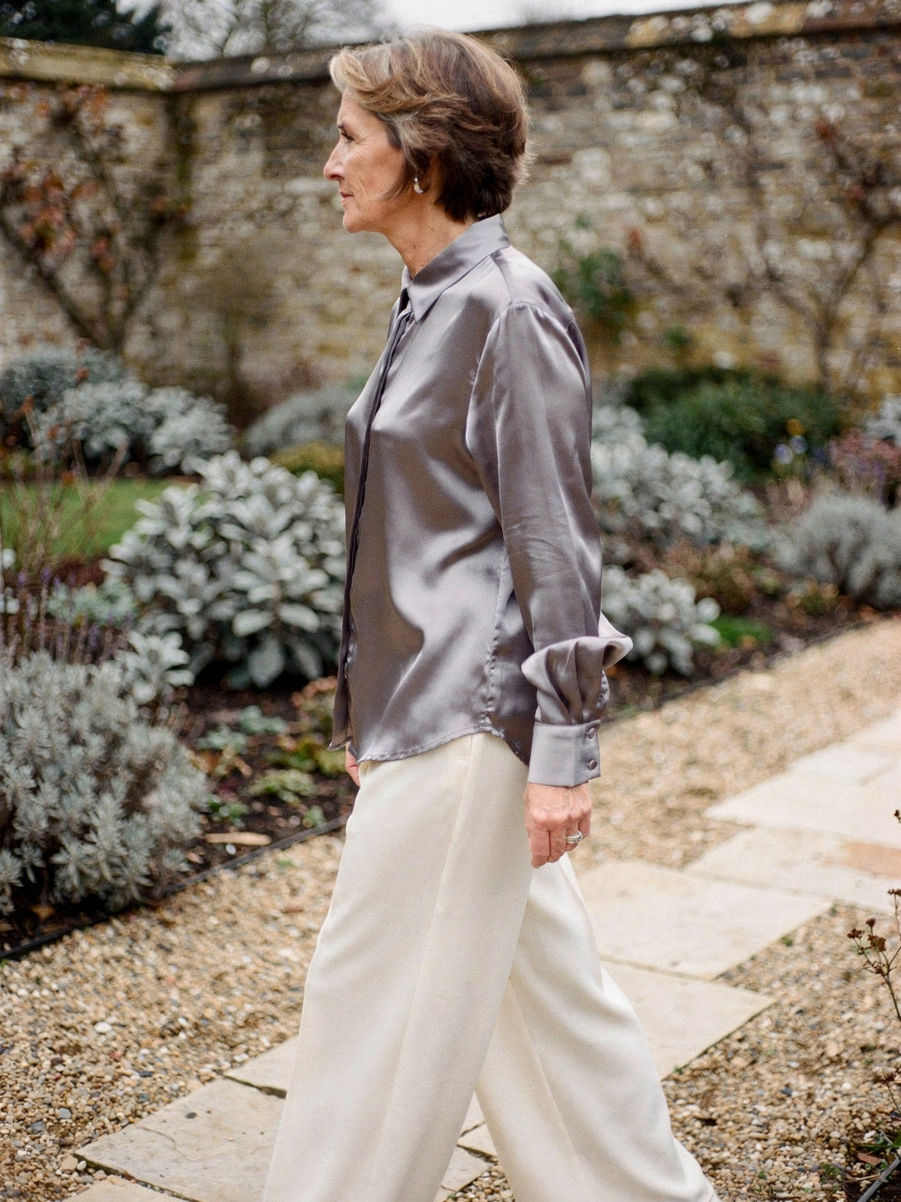 Woman wearing a pearl grey silk shirt over cream wide leg trousers in a walled garden