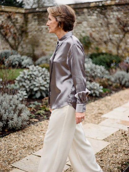 Woman wearing a pearl grey silk shirt over cream wide leg trousers in a walled garden