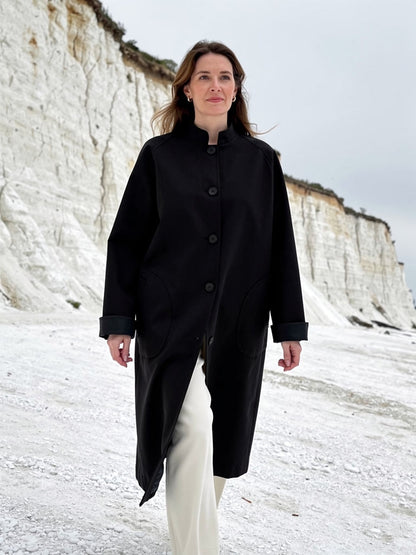 Womens stand collar coat in Blackthorn black, longline cocoon cut, worn against a white chalk cliff