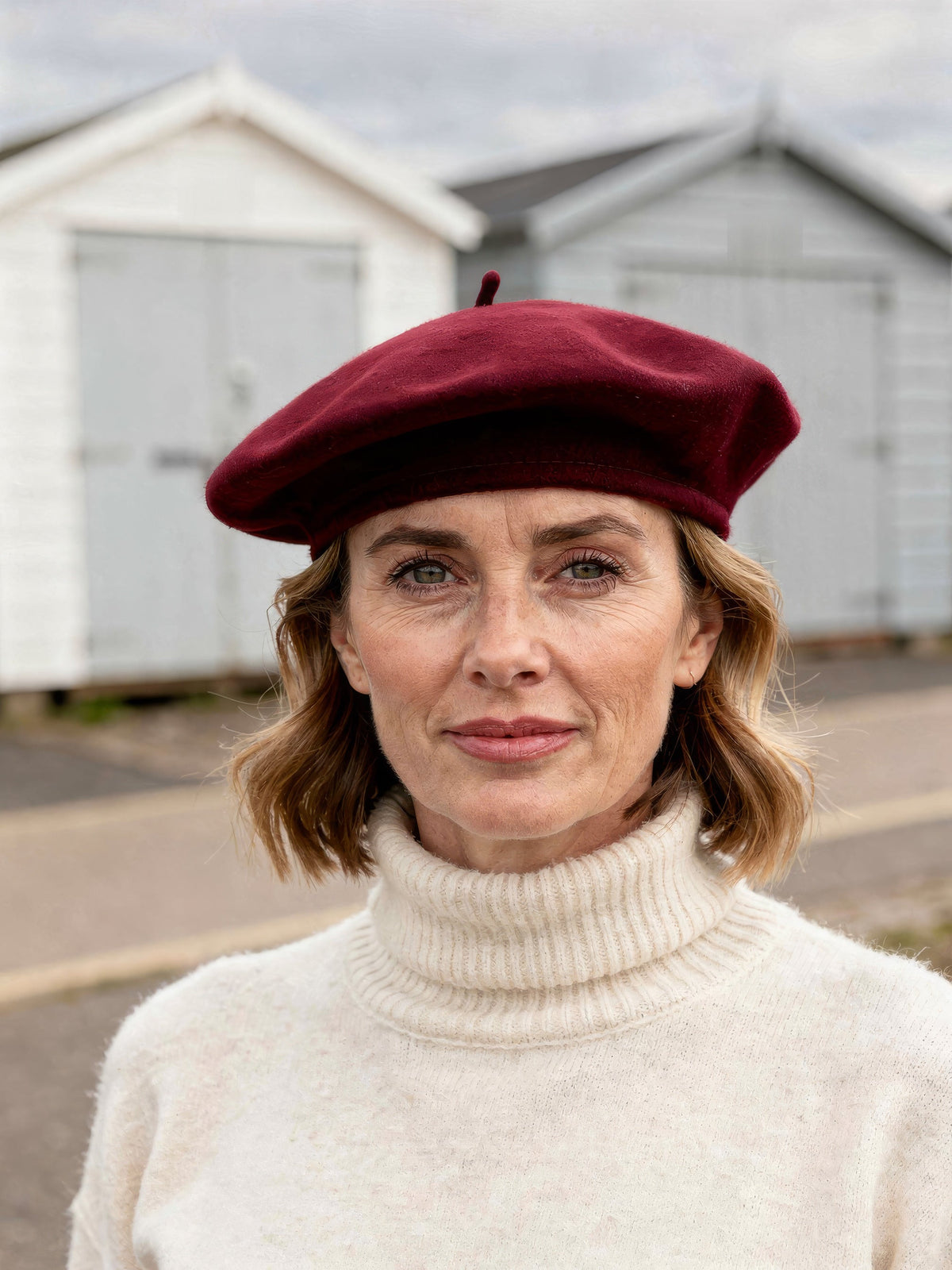 Womens wine red felt beret hat worn tilted back, seaside beach huts behind