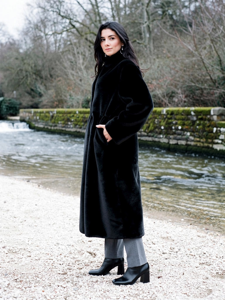 Woman wearing a black fluffy coat closed to the stand collar, standing on the gravel bank of a village weir