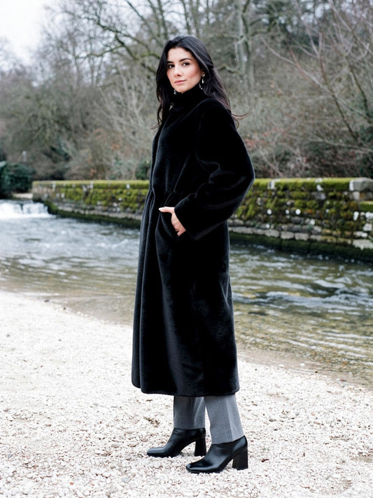 Woman wearing a black fluffy coat closed to the stand collar, standing on the gravel bank of a village weir