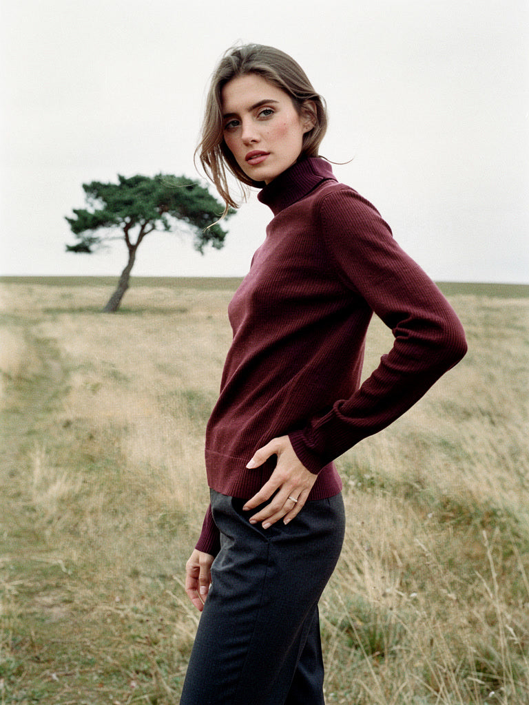 Woman on open chalk downland wearing a burgundy wool roll neck jumper, close-fitting fine rib with a folded roll neck