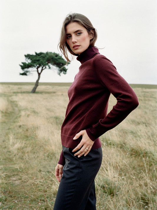 Woman on open chalk downland wearing a burgundy wool roll neck jumper, close-fitting fine rib with a folded roll neck