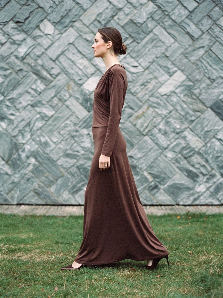 Wrap dress seen in profile, the A line skirt swinging as she walks past a herringbone slate wall